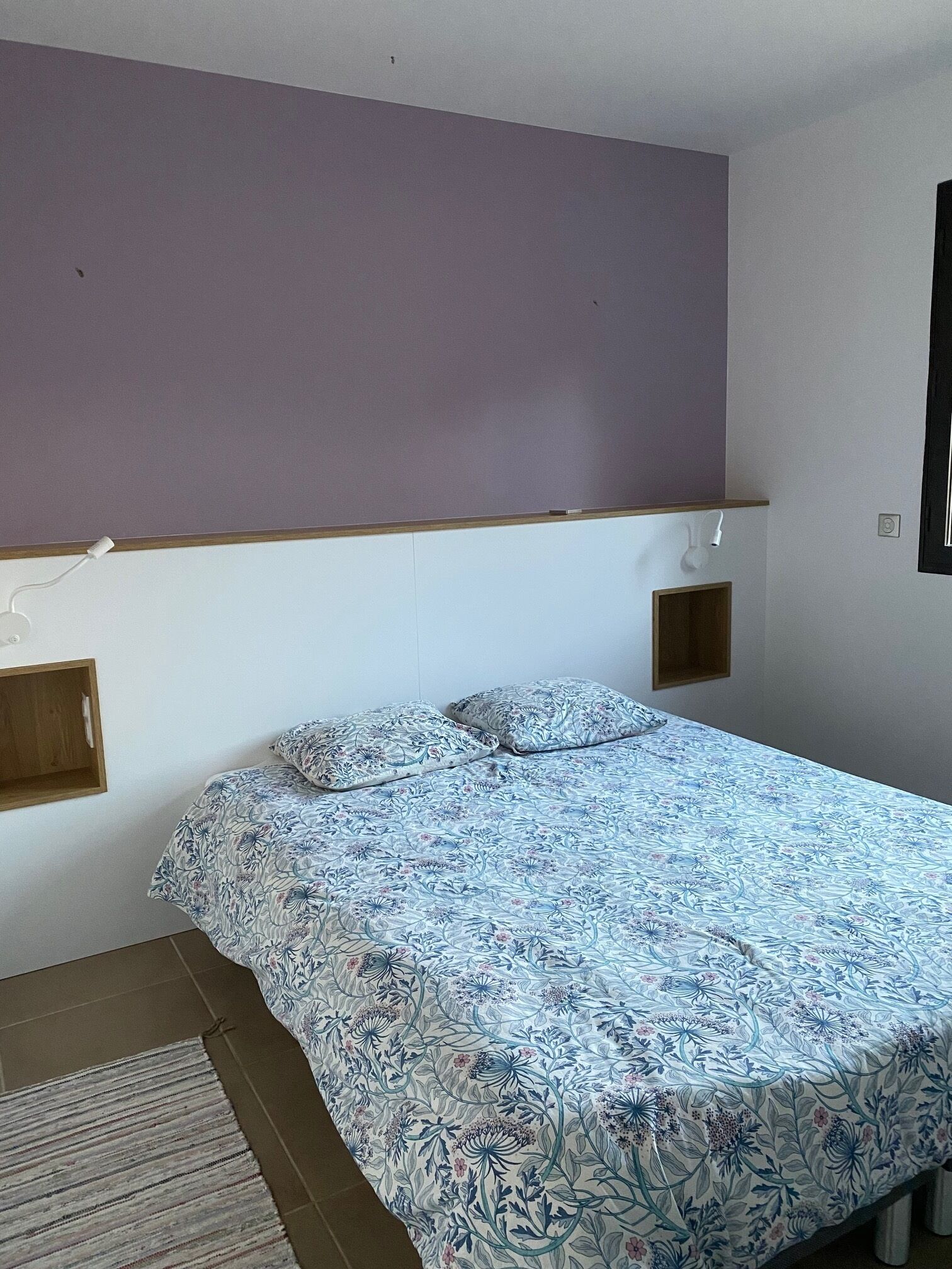 A bedroom with a bed, a nightstand, a wall-mounted shelf, and a window.