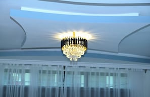 A chandelier with multiple lights hanging from the ceiling in a room with large windows and sheer curtains. - Lantana Hotel Kitui (Kitui)