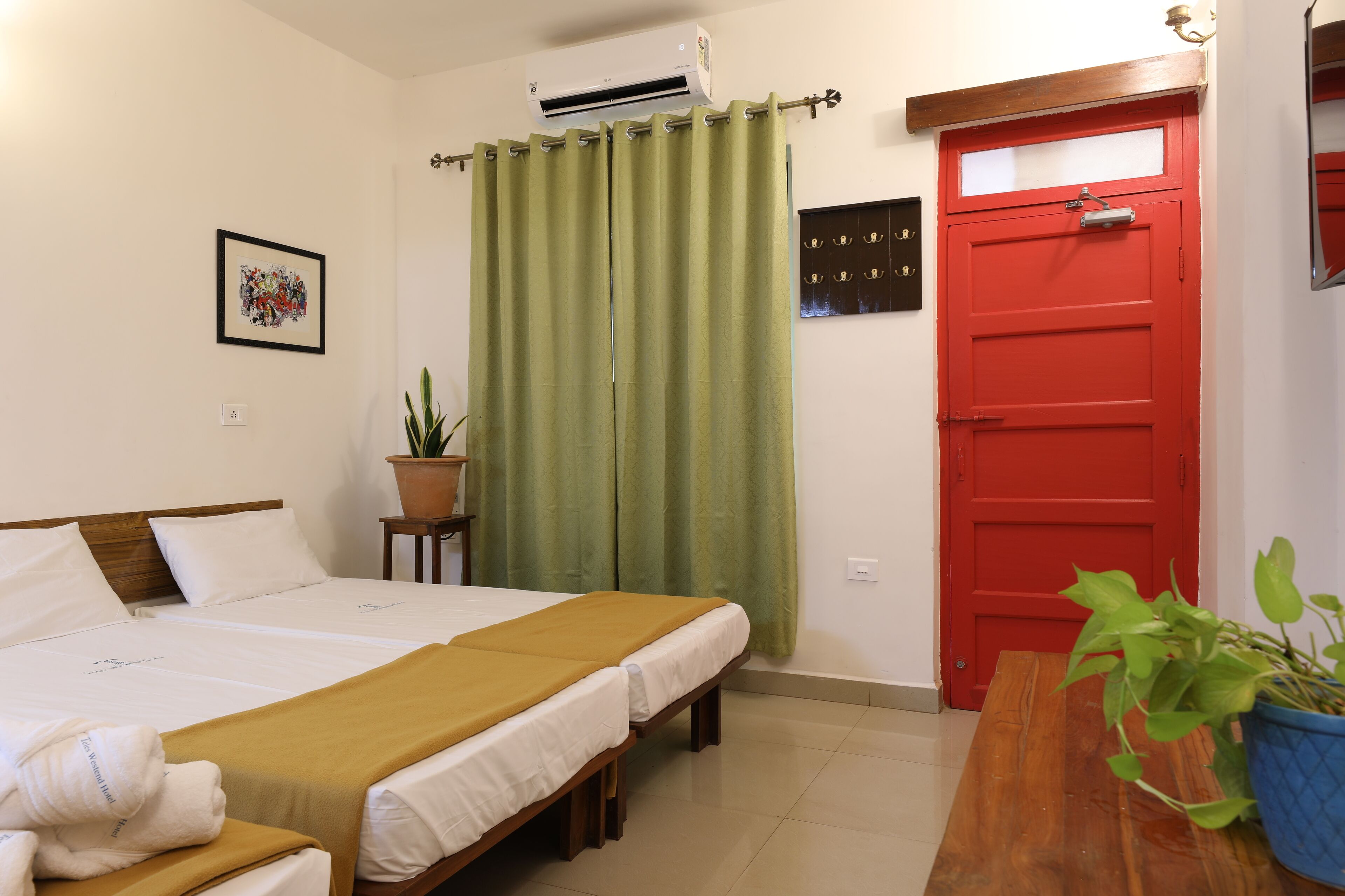 Habitación de hotel con cama, puerta roja, cortina verde, planta en maceta y un cuadro en la pared.
