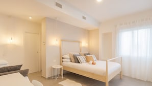 Une chambre à coucher avec un lit, une table de chevet et un petit tabouret. Il y a une fenêtre avec des rideaux et une porte.