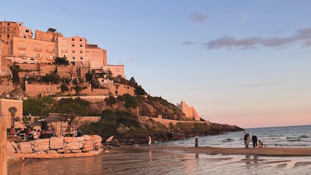 Une ville côtière avec des bâtiments historiques situés sur une colline, une plage avec des rochers et quelques personnes, et un ciel dégagé.