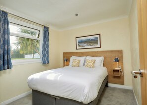 Une chambre à coucher avec un lit, une fenêtre avec des rideaux, une tête de lit, une table de chevet avec une lampe et un tableau encadré au mur.