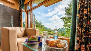 A cozy room with a sofa, a glass table with snacks, a bottle, and a view of greenery. - SRICHAN RESORTS (Pahalgam)
