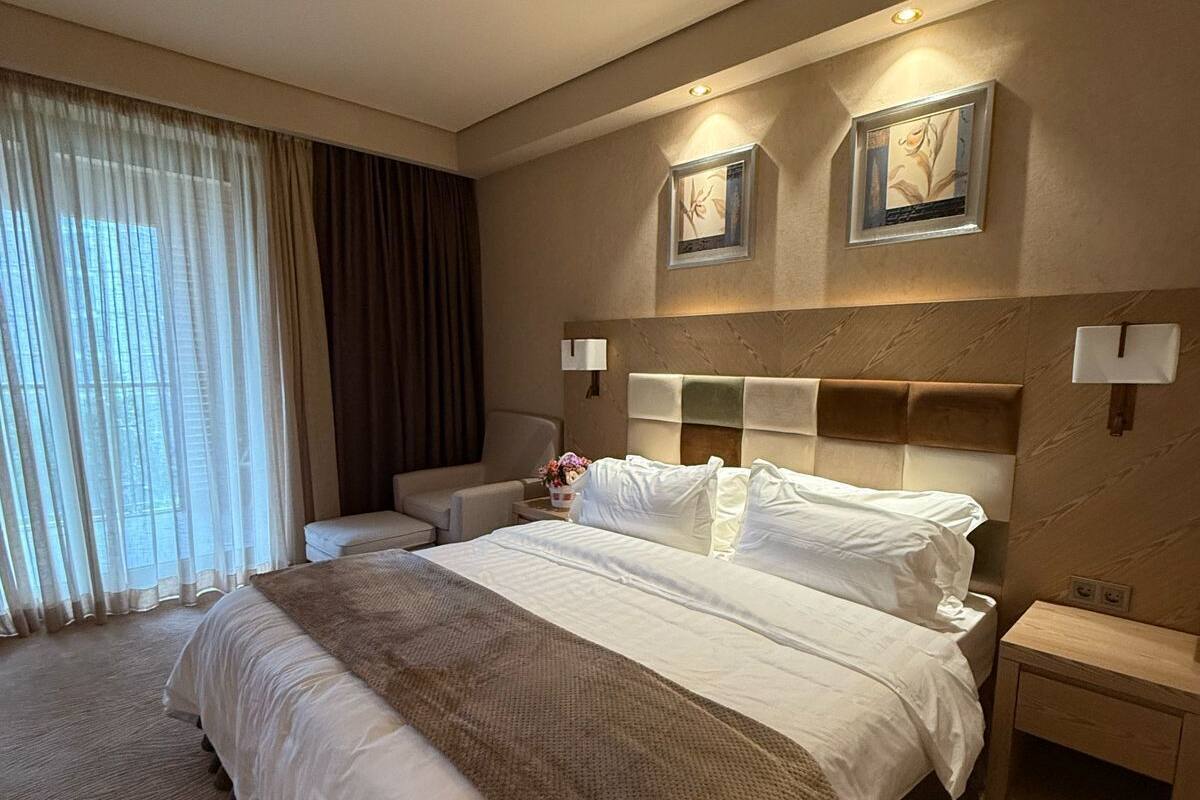 A hotel room with a large bed, a bedside table, a nightstand, a lamp, and a window with curtains.