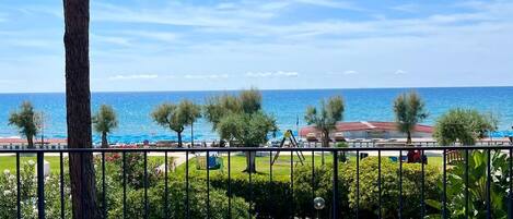 Un balcón con vista al mar, un árbol y un parque infantil al fondo.