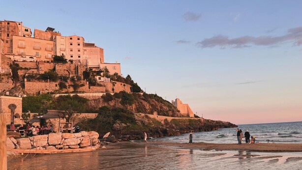 Une ville côtière avec des bâtiments historiques situés sur une colline, une plage avec des rochers et quelques personnes, et un ciel dégagé.