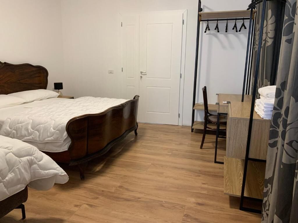 Een slaapkamer met een houten bed, een klein bureau en een stoel. Er is een kledingkast met daarin hangende kleding en een deur op de achtergrond.