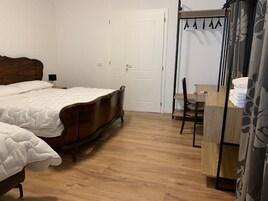 A bedroom with a wooden bed, a small desk, and a chair. There is a wardrobe with hanging clothes and a door in the background.