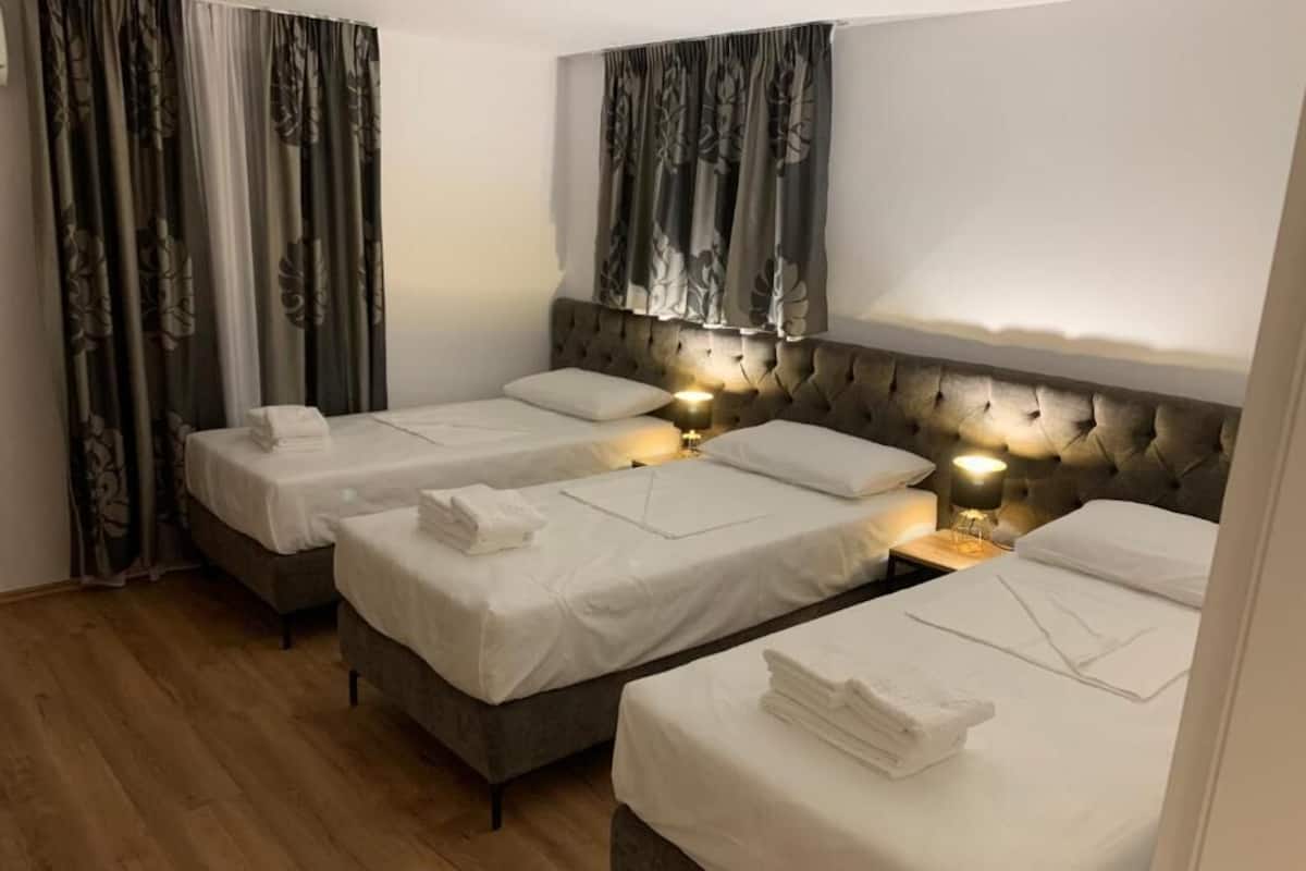 A hotel room with three beds, each with white linens and a neatly folded towel. The room has a wooden floor and a headboard with a tufted design.