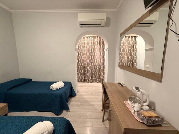 A hotel room with two single beds, a wooden desk with a telephone and a kettle, and an air conditioning unit on the wall.