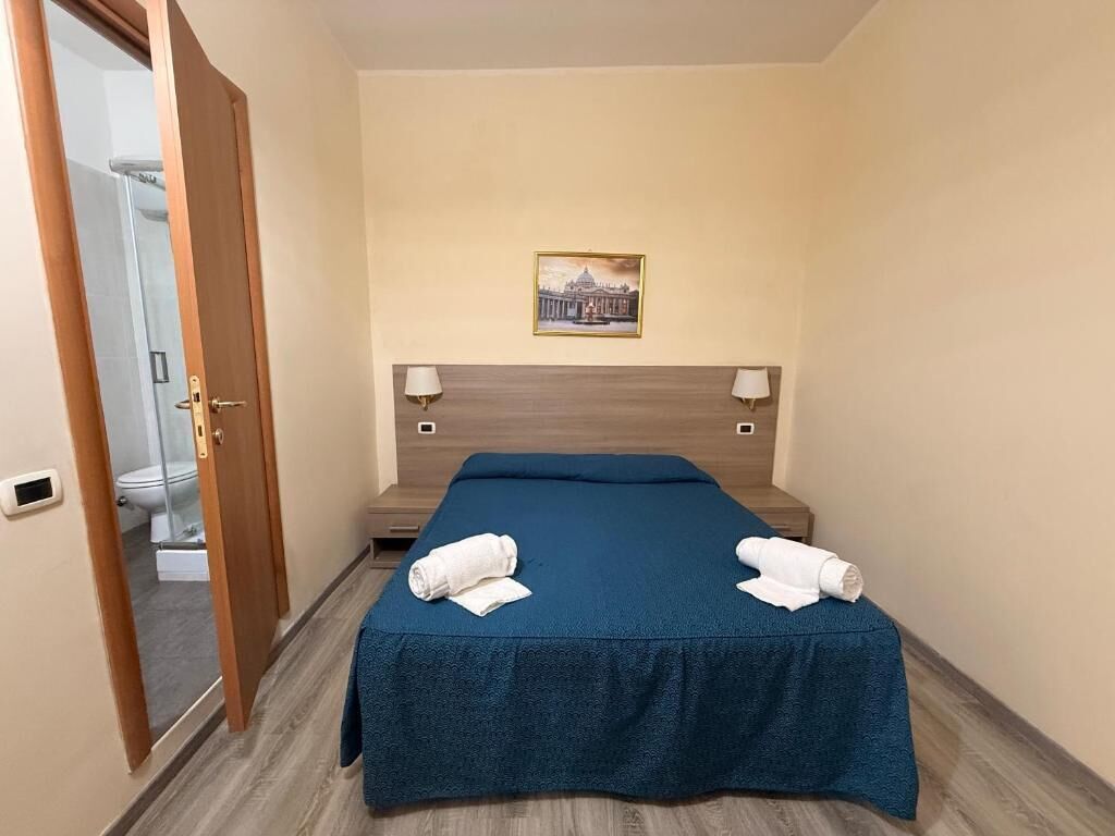 A hotel room with a bed, two bedside lamps, a framed picture on the wall, and a door leading to a bathroom.