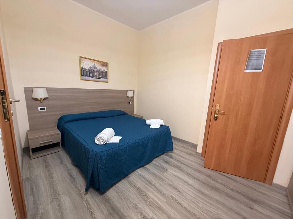 A hotel room with a bed, a nightstand, a lamp, a door, and a framed picture on the wall.