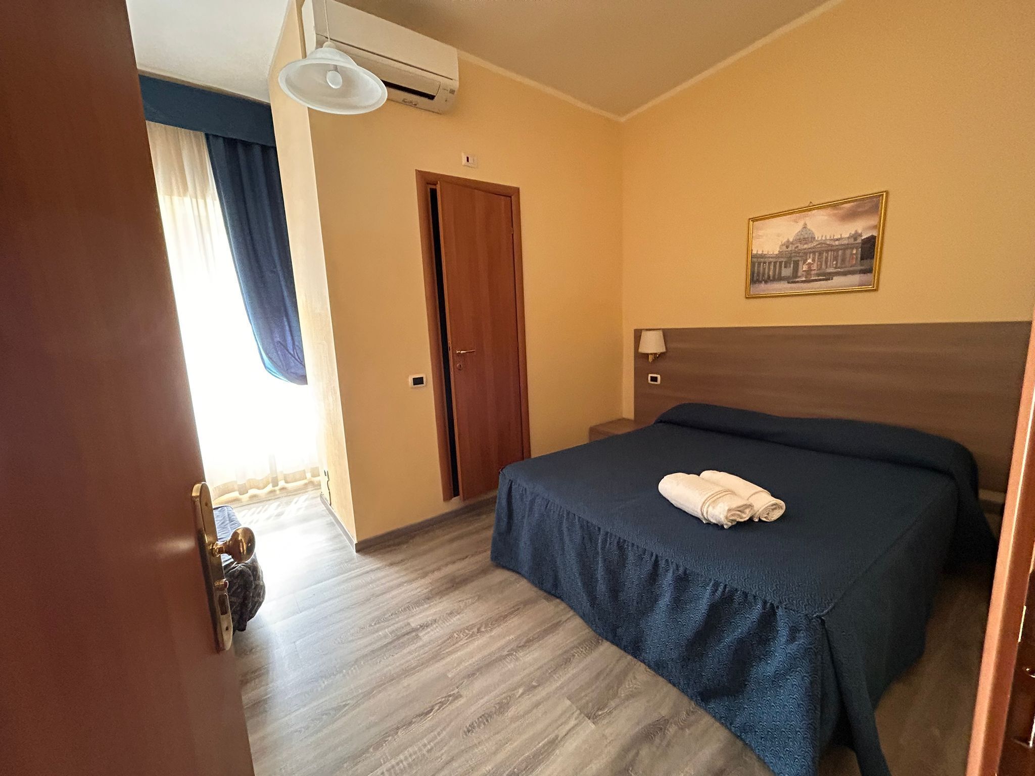 Una habitación de hotel con cama, puerta, un cuadro y un equipo de aire acondicionado.