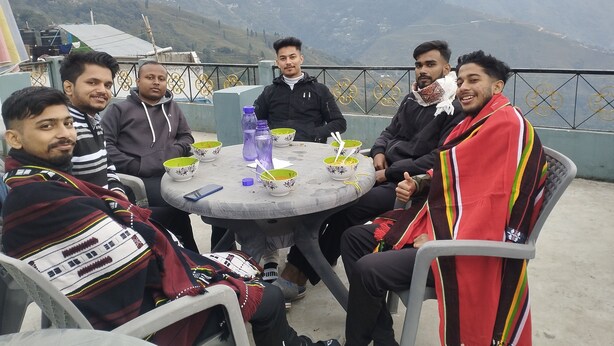 Een groep mensen zit aan een ronde tafel buiten, met een prachtig uitzicht op de bergen op de achtergrond.