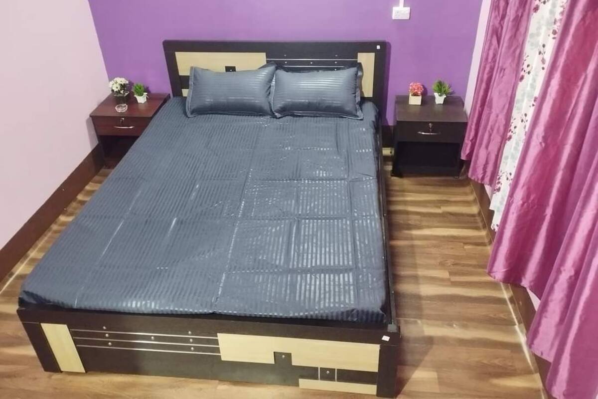 Una cama con colchón y cabecera azules, dos mesitas de noche con adornos, suelo de madera y pared morada.