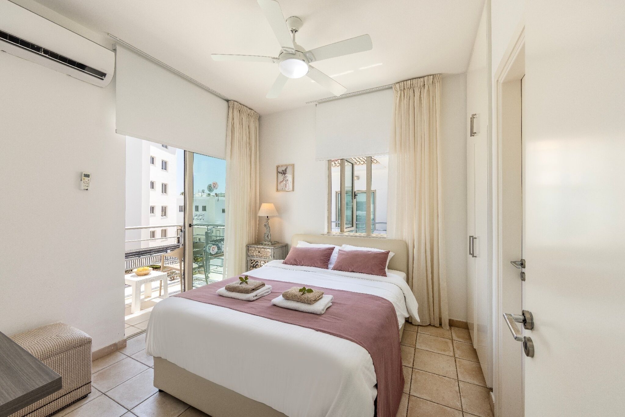 Quarto de hotel com cama, ventilador de teto, janela com cortinas e vista para edifícios.