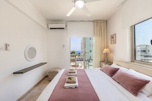 A hotel room with a bed, a ceiling fan, a window with a view, a bedside table with a lamp, and a wall-mounted air conditioner. - 4 bedroom villa by Fig tree bay beach (Protaras)