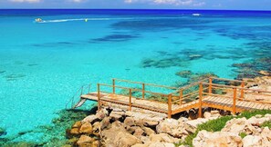 A wooden pier extending over clear turquoise waters with rocky outcrops, leading to a small island. - 4 bedroom villa by Fig tree bay beach (Protaras)