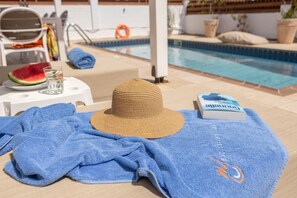 A poolside area with a sun lounger, a hat, a towel, a glass of water, a watermelon slice, and a book. - 4 bedroom villa by Fig tree bay beach (Protaras)