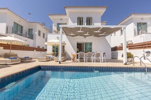 A swimming pool with a tiled edge, a covered patio area with outdoor furniture, and a white building with balconies. - 4 bedroom villa by Fig tree bay beach (Protaras)