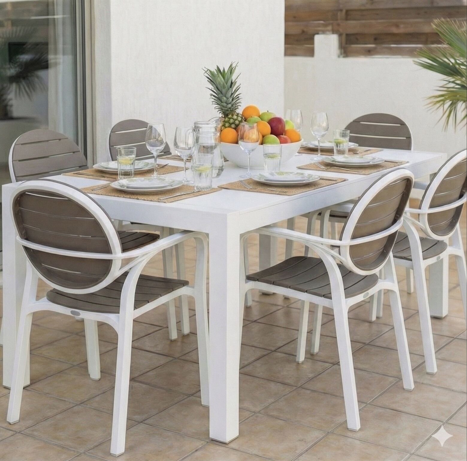 A white dining table with four chairs, a fruit bowl, and glasses on a tiled patio.
