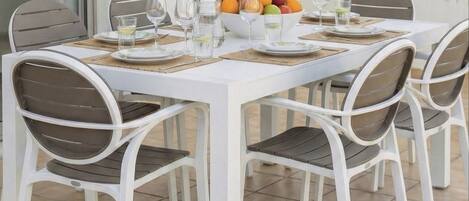 A white dining table with four chairs, a fruit bowl, and glasses on a tiled patio.