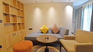 A modern living room with a sofa, armchair, ottoman, and a coffee table. There is a wooden shelving unit and a large window with curtains. - Hotel Sands Point (Puducherry)