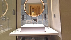 A bathroom with a white sink, a round mirror, and a shower. - Hotel Sands Point (Puducherry)
