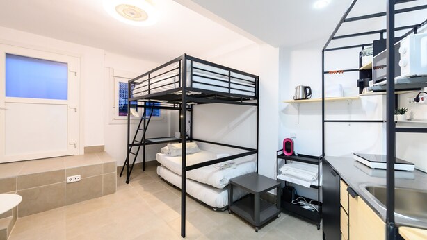 Une chambre avec un lit superposé, un coin cuisine, une fenêtre avec des stores et une petite table.