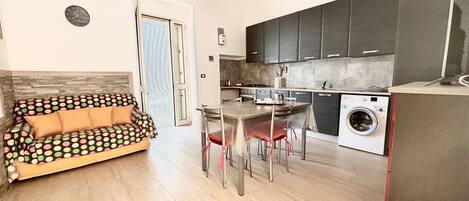 En kompakt opholdsstue med sofa, spisebord, stole, vaskemaskine og mørke skabe.