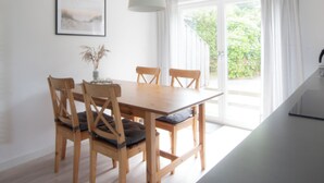 Et spiseområde med et træbord og stole, en hængende lampe, et indrammet billede på væggen og udsigt gennem en glasdør udendørs.