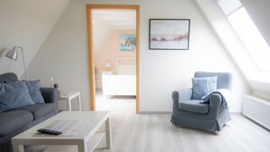 En moderne stue med sofa, lænestol og sofabord. Der er en dør, der fører til et andet rum, og et billede i ramme på væggen.