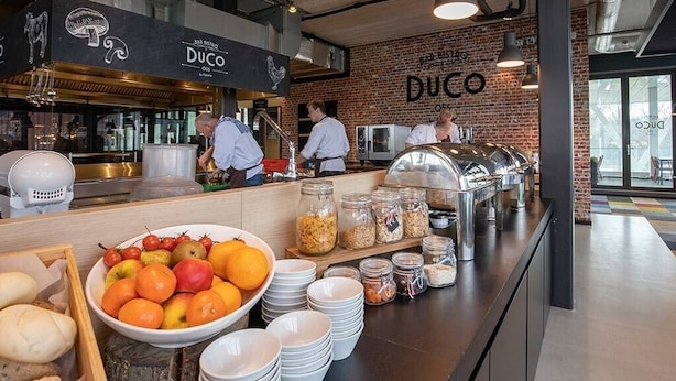 Een ontbijtbuffet met diverse soorten eten, zoals fruit, ontbijtgranen en brood, in een moderne setting met zichtbare bakstenen muren en verlichting in industriële stijl.