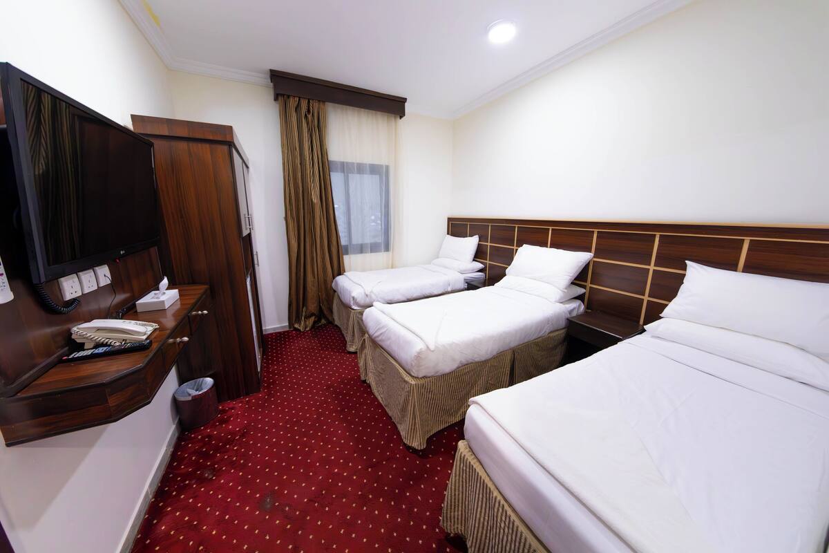 Ein Hotelzimmer mit zwei Einzelbetten, einem hölzernen Kopfteil, einem kleinen Schreibtisch mit Telefon und Lampe sowie einem roten Teppichboden.