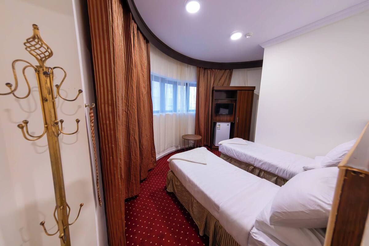 Ein Hotelzimmer mit zwei Betten, einem kleinen Tisch, einem Kleiderständer und einem Fenster mit Vorhängen.