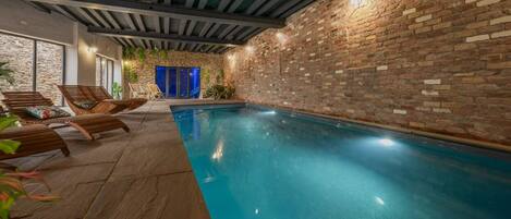 An indoor swimming pool with a brick wall, wooden lounge chairs, and a stone floor.