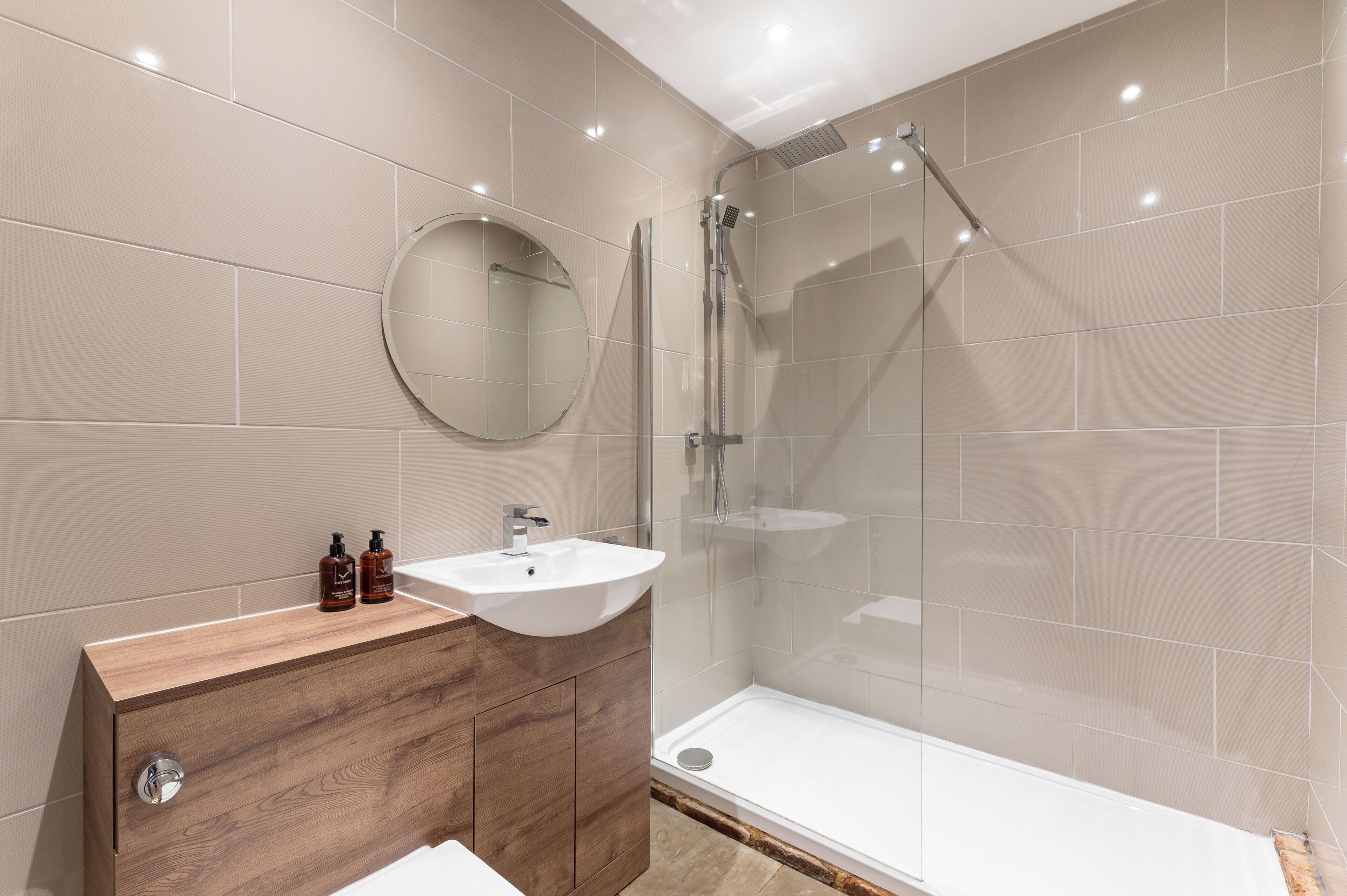 A modern bathroom with a wooden vanity, a round mirror, a white sink, and a glass-enclosed shower.