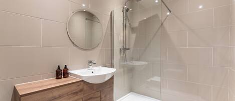 A modern bathroom with a wooden vanity, a round mirror, a white sink, and a glass-enclosed shower.