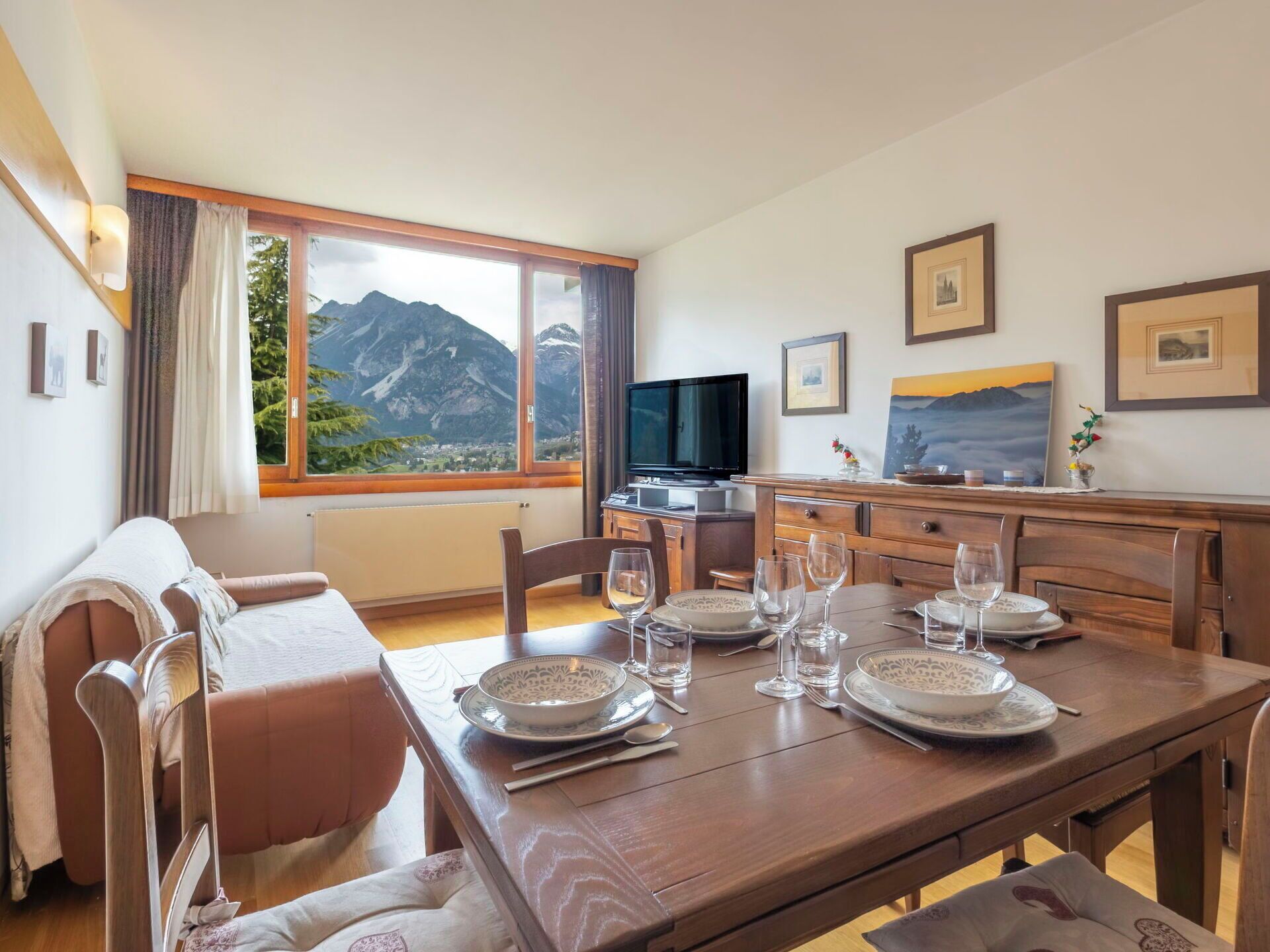 Uma sala de jantar com mesa posta para quatro pessoas, uma televisão e vista para as montanhas.