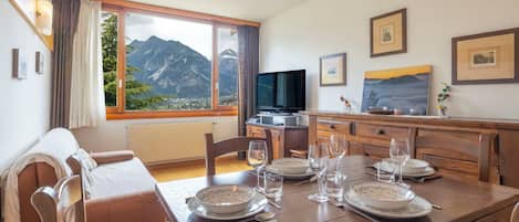 Uma sala de jantar com mesa posta para quatro pessoas, uma televisão e vista para as montanhas.