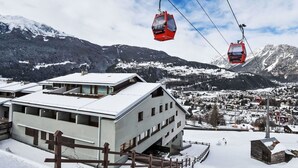 En modern byggnad med ett platt tak, ett trästaket och en liten träkonstruktion i ett snöigt landskap med berg i bakgrunden.
