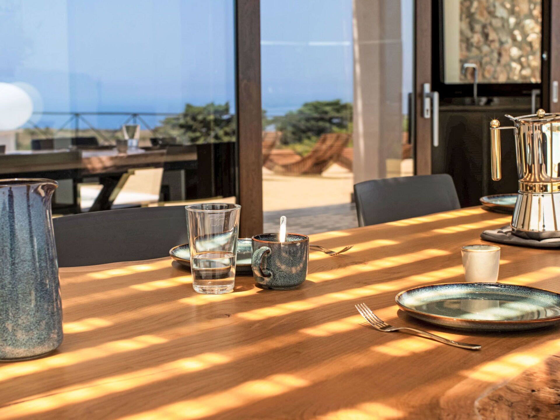 Une table à manger baignée de lumière, avec de beaux verres transparents, une cafetière et une vue sur l’extérieur.