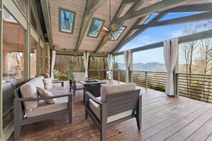 A covered outdoor seating area with a wooden deck, cushioned seating, and a view of the mountains. - Astonishing Views, Gas Fire Table, Outdoor Living (Waynesville)