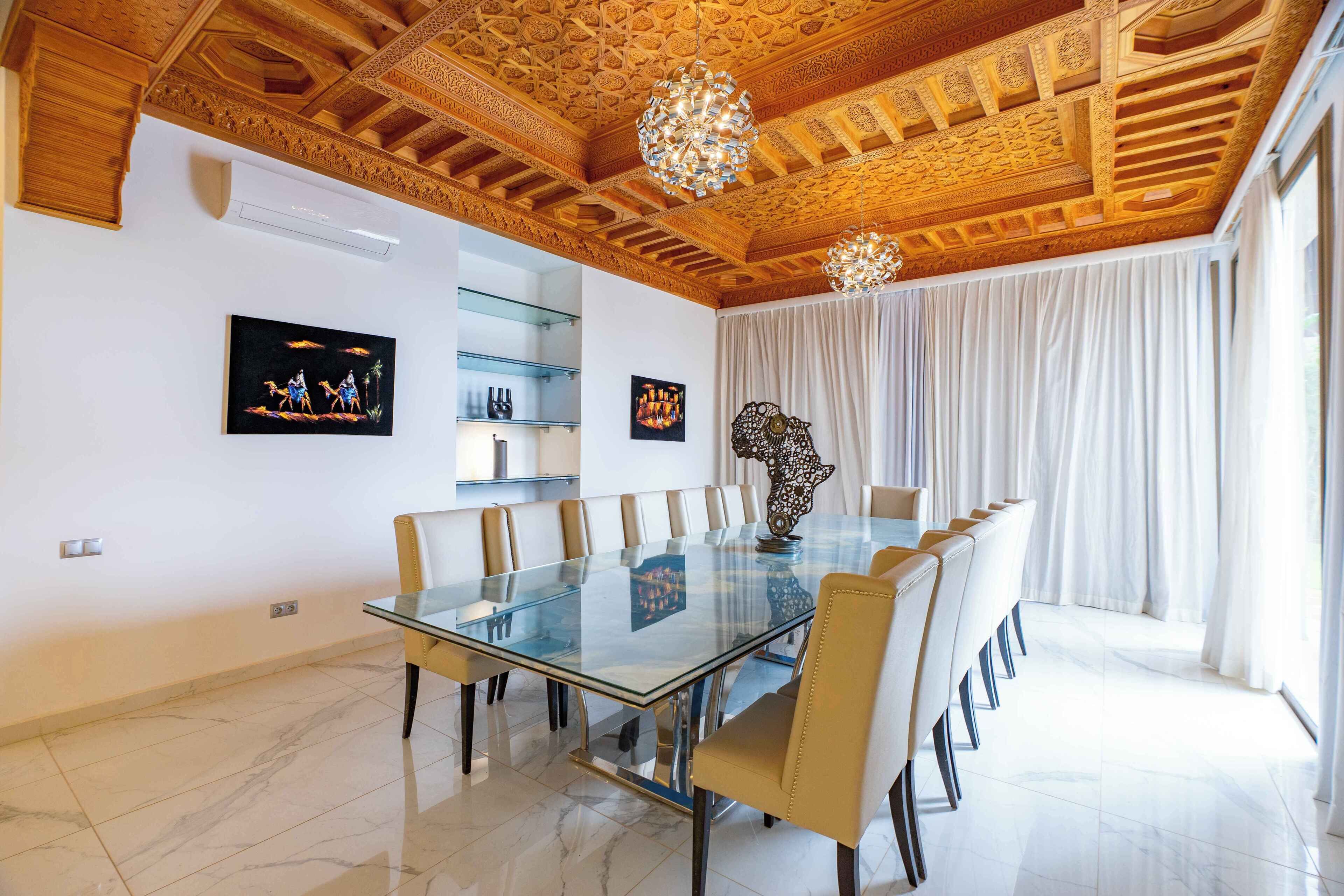 A spacious dining room with a glass table surrounded by chairs, ornate wooden ceiling, and large windows with curtains.