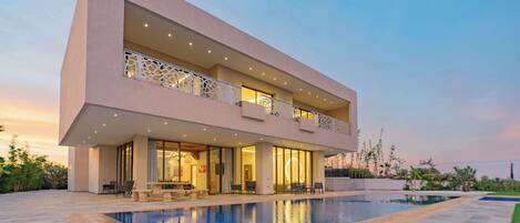 A modern two-story house with a swimming pool, outdoor seating, and a well-lit balcony.