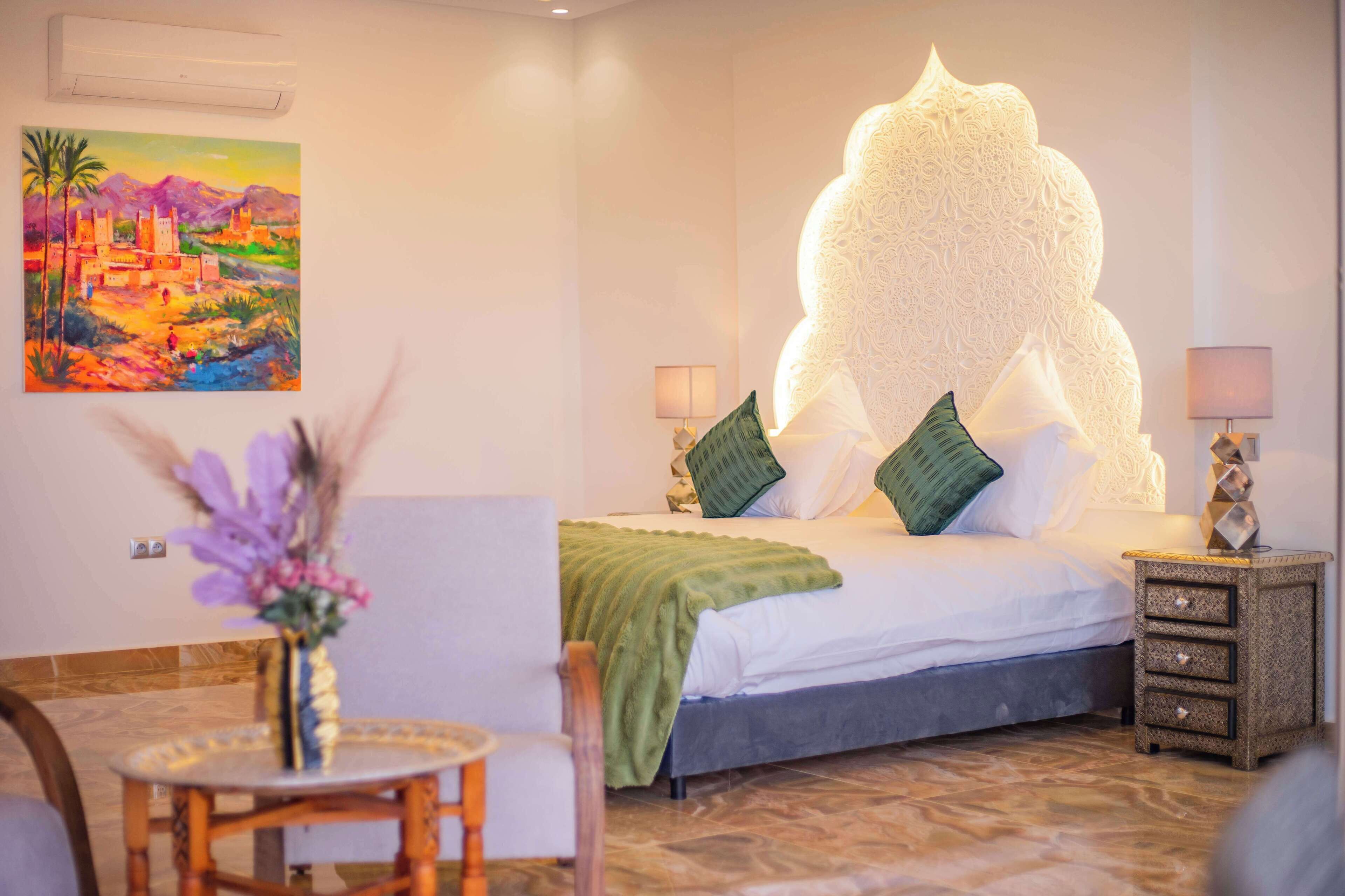 A hotel room with a bed, a nightstand, a lamp, a chair, a table, and a painting on the wall.