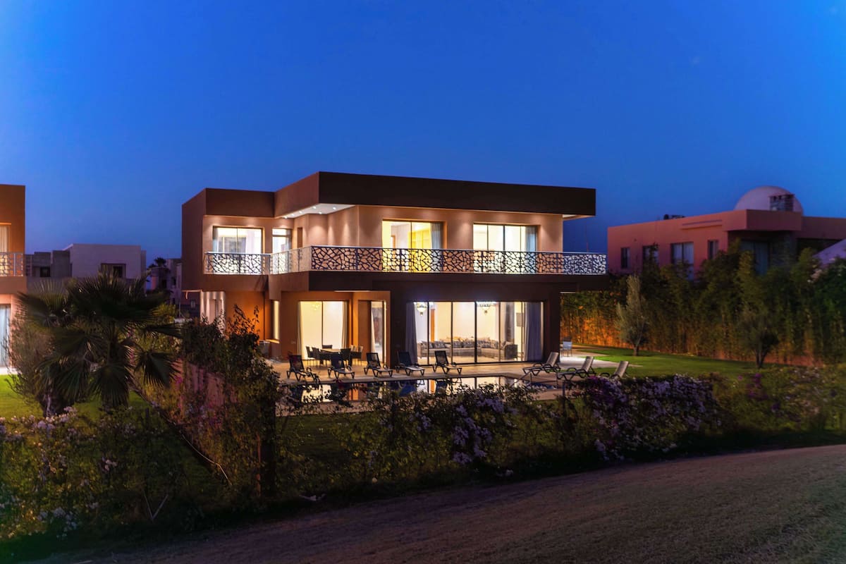 A modern two-story house with a balcony, surrounded by lush greenery and a well-maintained lawn.