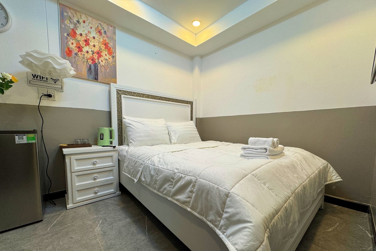 A hotel room with a bed, a nightstand, a kettle, a refrigerator, a painting, and a sign indicating WiFi password availability.