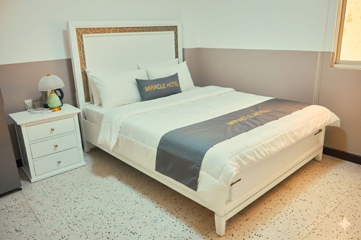 A neatly arranged hotel room with a bed, a bedside table with a lamp, and a small drawer unit.