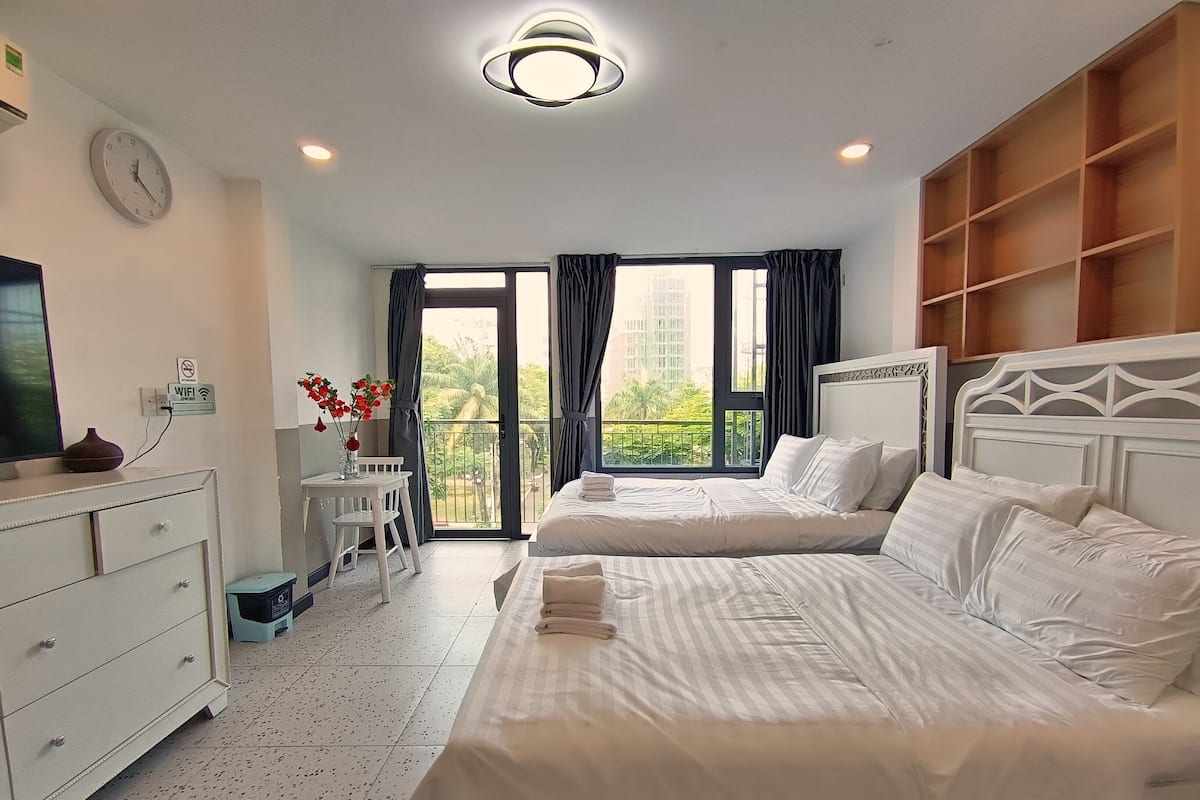 A hotel room with two beds, a desk, a dresser, a TV, and a balcony with curtains.